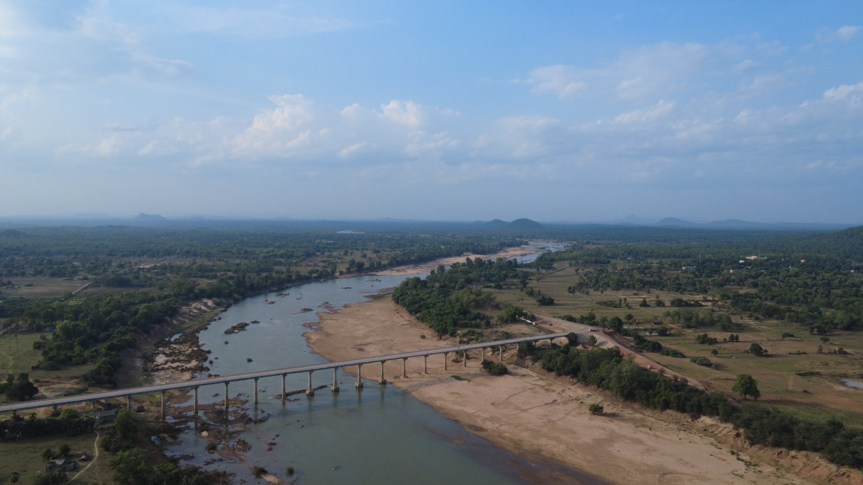 A long concrete bridge spanning a wide, shallow river in a vast, green landscape.